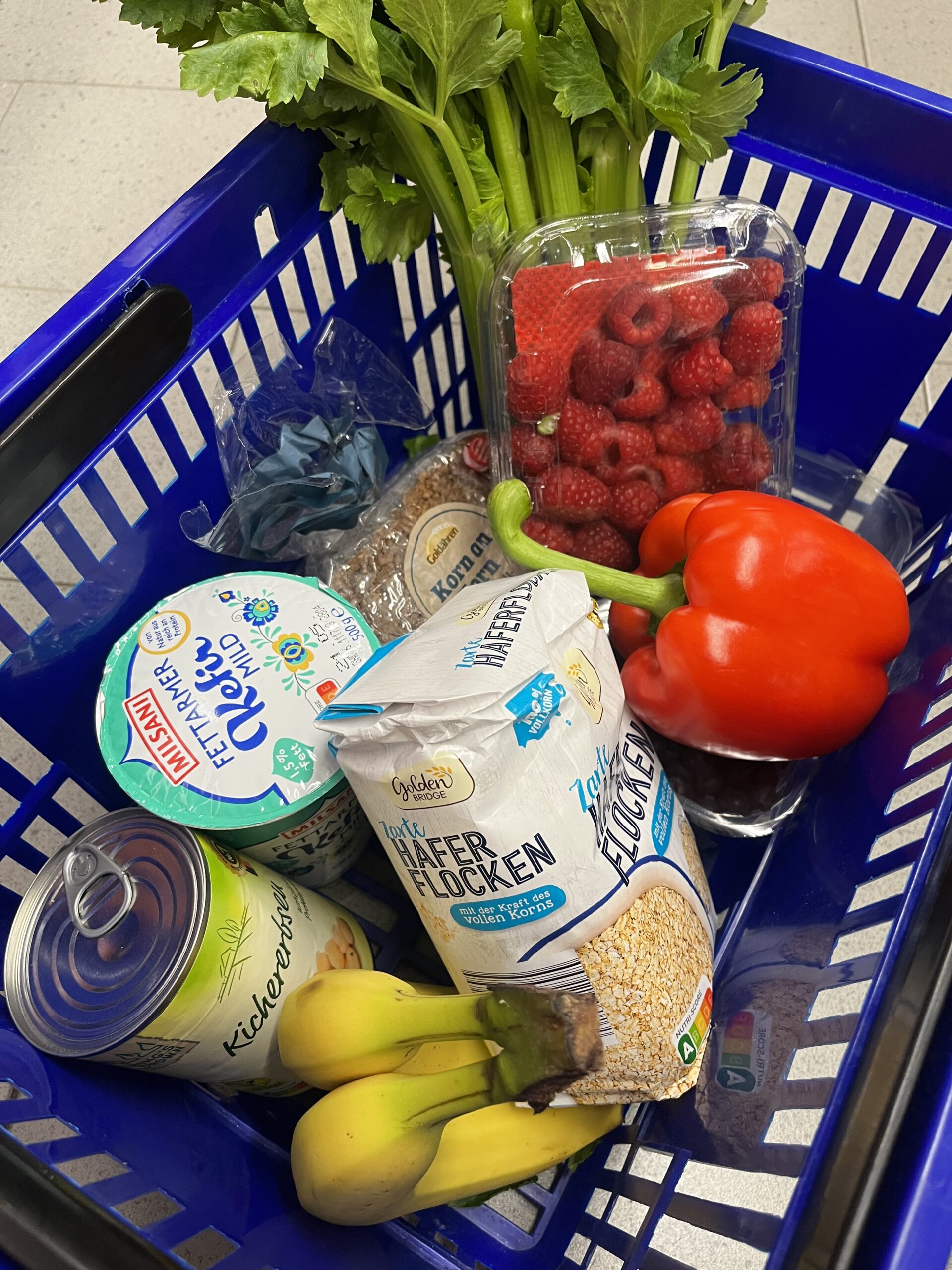 Ein blauer Einkaufskorb gefüllt mit darmfreundlichen Lebensmitteln: Sellerie, Himbeeren, rote Paprika, natürliche Haferflocken, ein Becher Kefir, eine Packung Kornbrot, 2 Bananen und eine Dose Kichererbsen.
